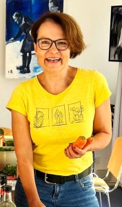 Petra Paumann, eine der Gastgeberinnen der Detox-Tage, hält eine Karotte in der Hand und lächelt. Sie trägt ein gelbes T-Shirt mit Pflanzenmotiven.